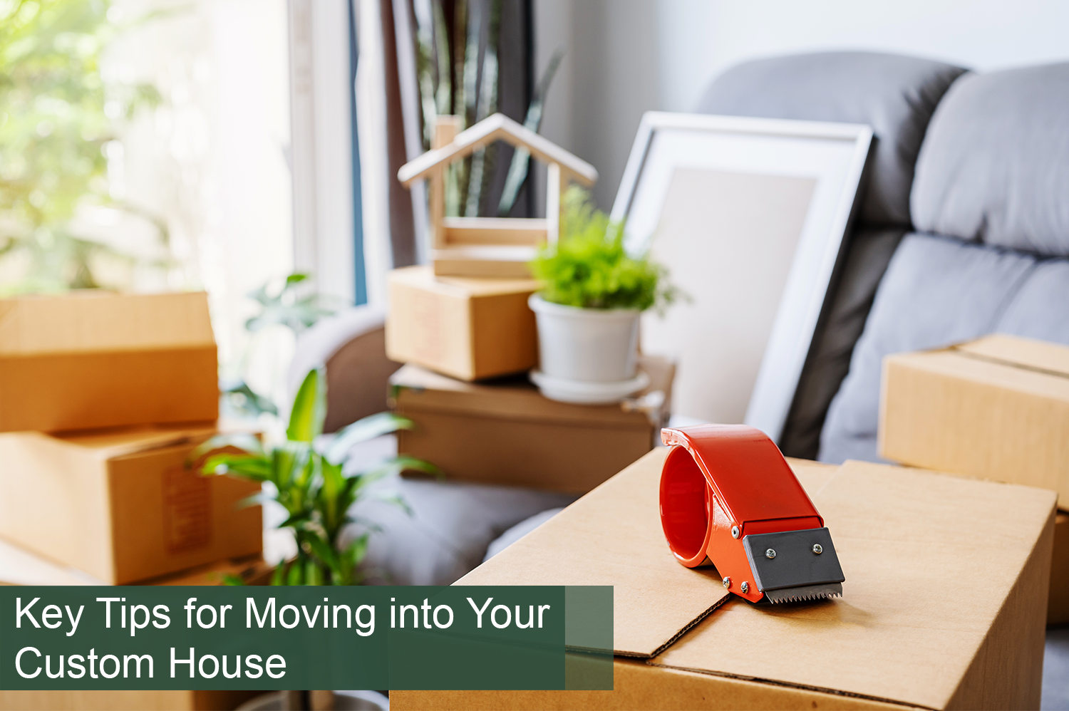 A home packed up with cardboard boxes, picture frames, and plants on a grey couch, ready to move into a custom house.